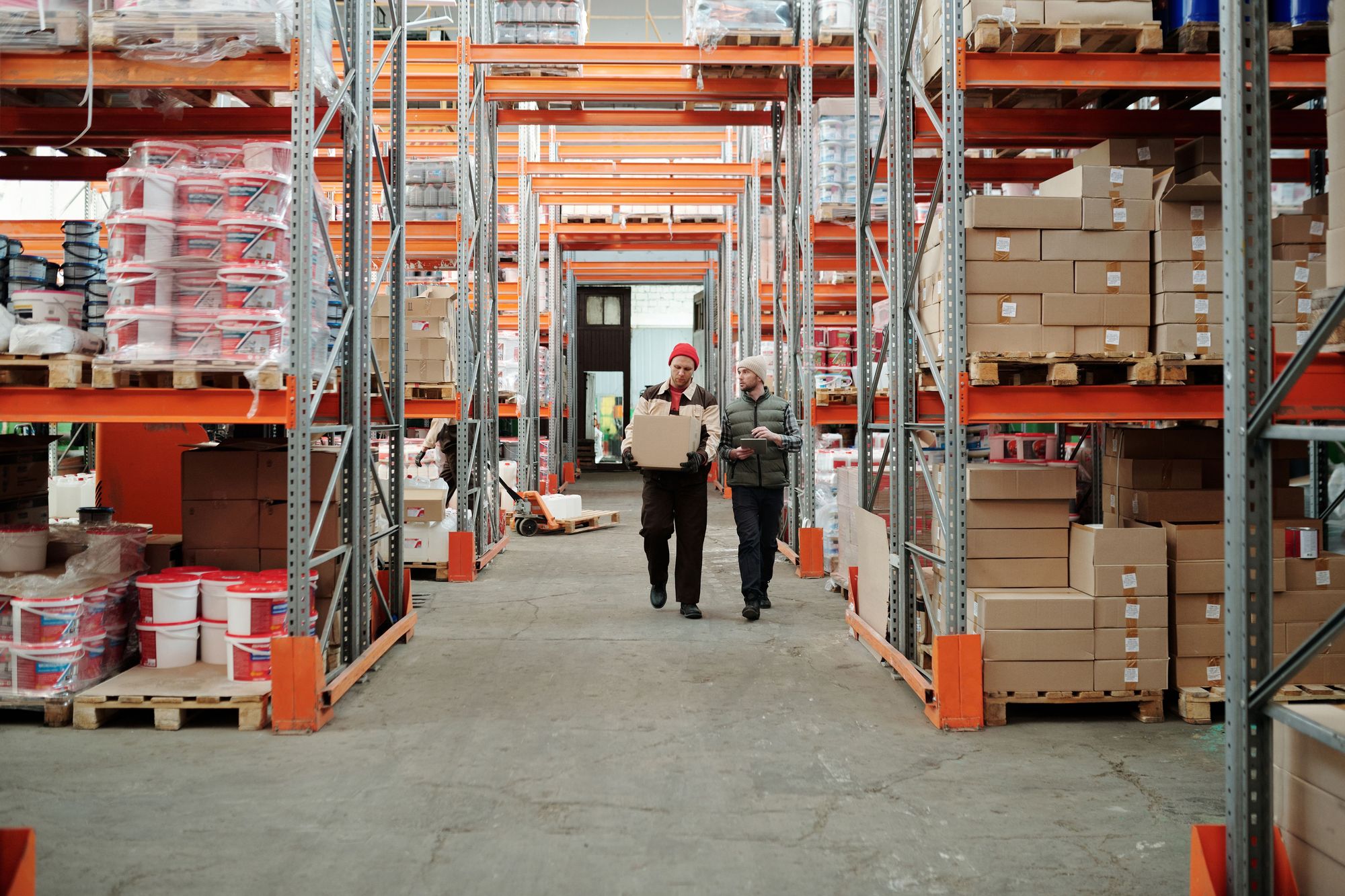 Two workers walking through a distribution warehouse stacked with pallets and cartons